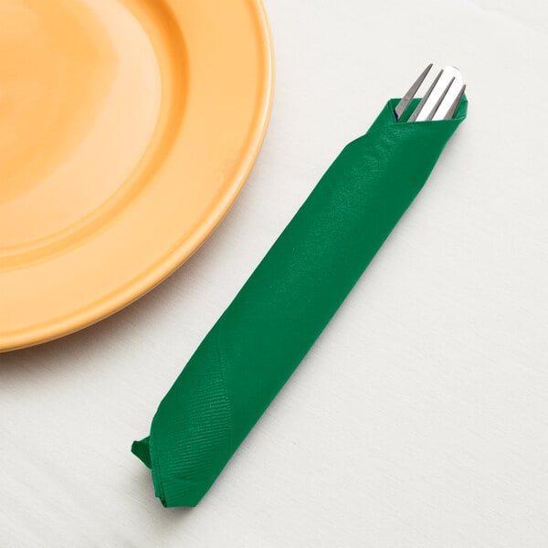 A fork and knife wrapped in an emerald green Creative Converting paper dinner napkin on a table next to a plate.