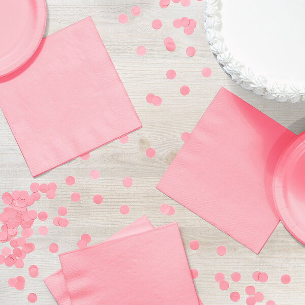 A table with a white cake and pink paper plates and napkins, with a pink napkin on the table with confetti.