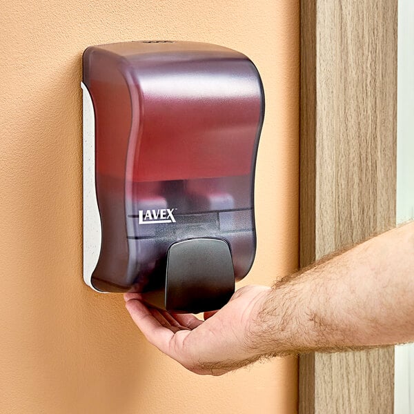 A hand using a Lavex manual foaming soap dispenser on a wall.