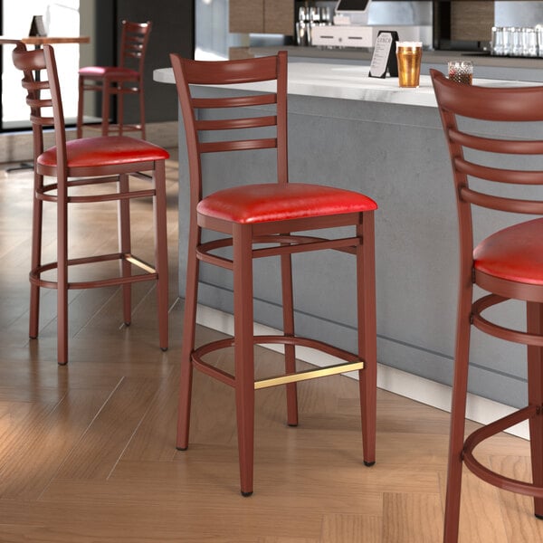 A Lancaster Table & Seating metal ladder back bar stool with a mahogany wood grain finish and red vinyl seat on a metal frame.