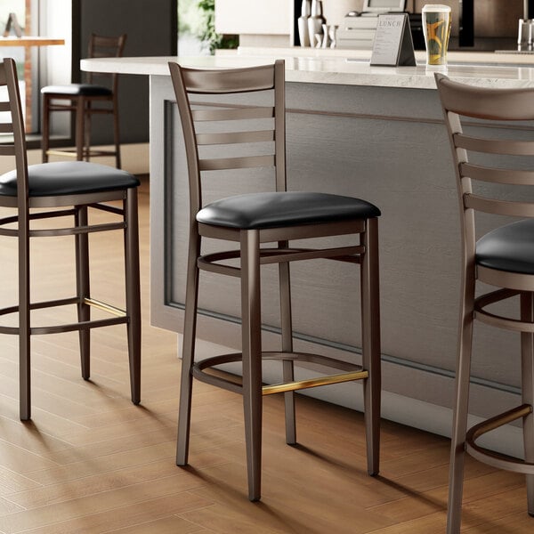 Three Lancaster Table & Seating Spartan Series Metal Ladder Back Bar Stools with Black Vinyl Seats on a counter.