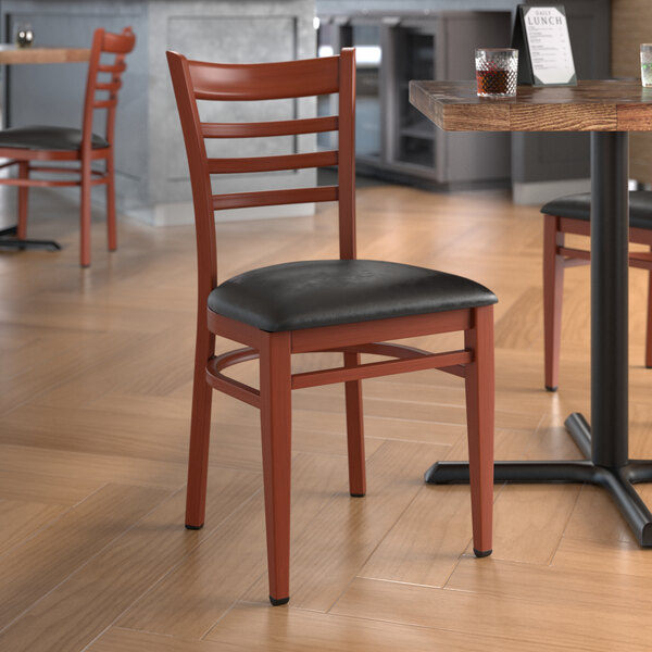 A Lancaster Table & Seating metal ladder back chair with mahogany wood grain finish and black vinyl seat at a table in a restaurant.