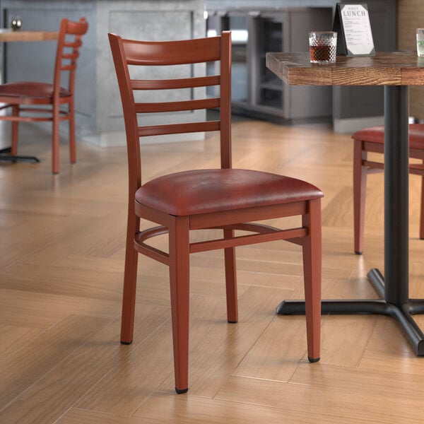 A Lancaster Table & Seating metal ladder back chair with mahogany wood grain finish and burgundy vinyl seat on a table in a restaurant.