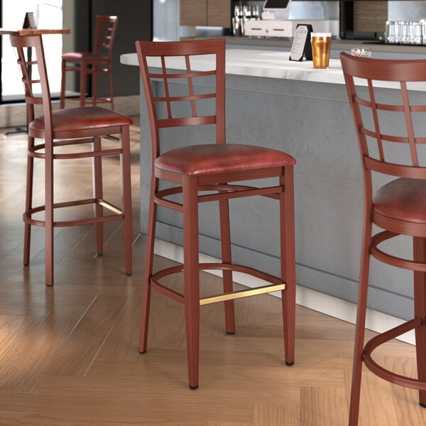 A Lancaster Table & Seating metal bar stool with mahogany wood grain finish and a burgundy vinyl seat.