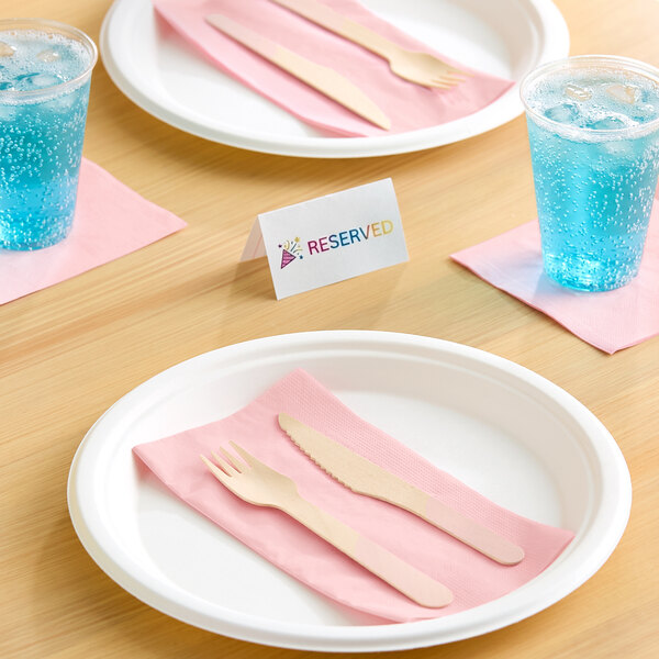 A table setting with white plates, pink napkins, wooden cutlery, clear cups with blue fizzy drinks, and a reserved sign.