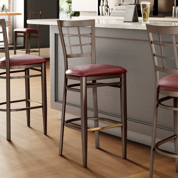 Three Lancaster Table & Seating metal bar stools with dark walnut wood grain and burgundy vinyl seats at a kitchen counter.