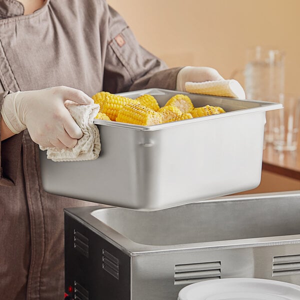 A woman in a chef's uniform holding a Vollrath stainless steel steam table pan of corn.