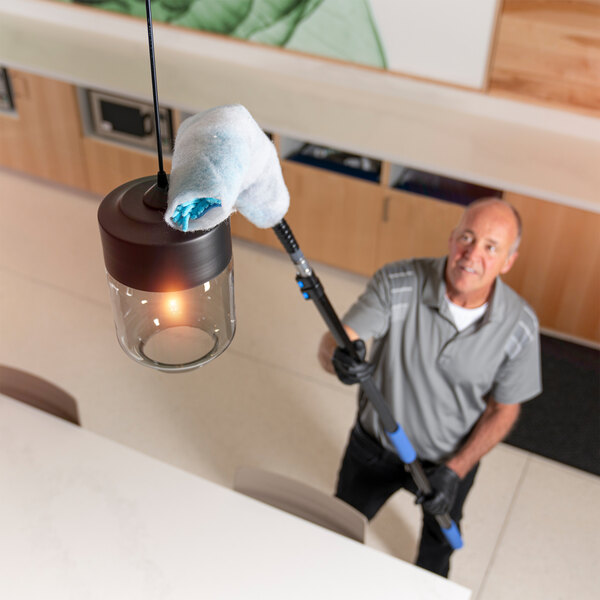 A person using a long-handled tool with a blue duster sheet to clean a hanging light fixture.