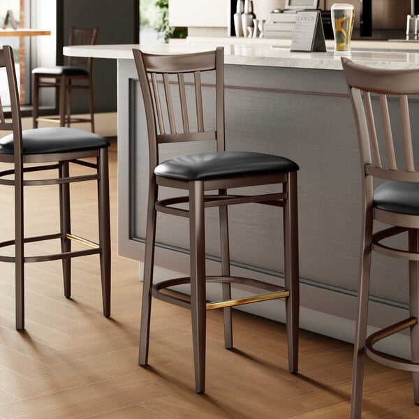 Three Lancaster Table & Seating Spartan metal slat back bar stools with black vinyl seats next to a counter.