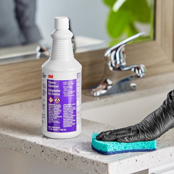 A person in black gloves cleaning a countertop with 3M Creme Cleanser using a sponge.