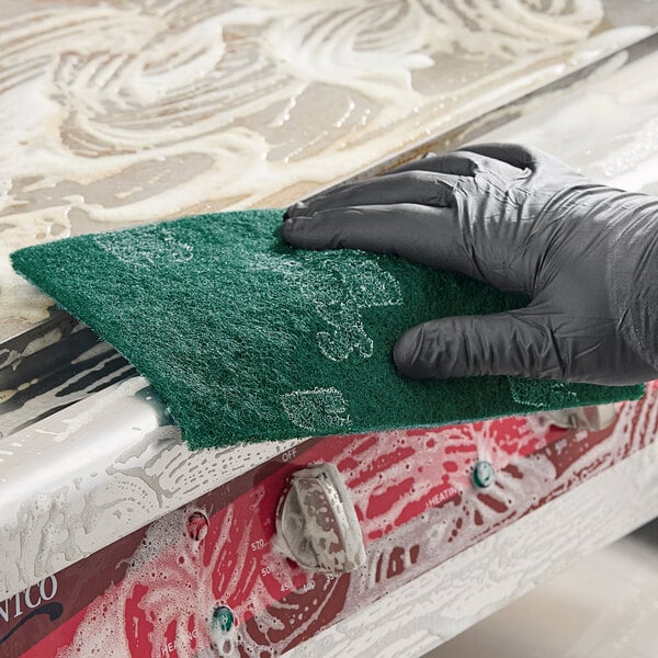 A green 3M Scotch-Brite general purpose scouring pad being used to clean a soapy metal surface.