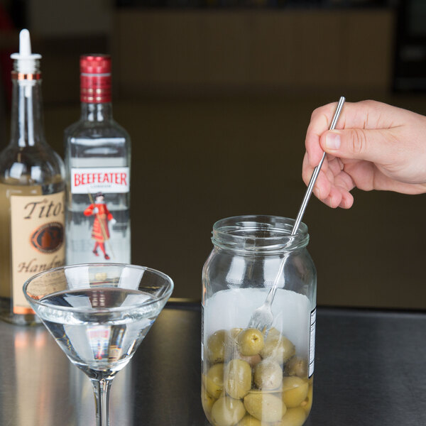A stainless steel American Metalcraft bar fork in a jar of green olives next to a martini glass on a counter.