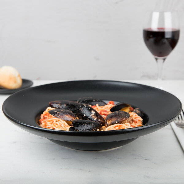 A black Hall China Mediterranean pasta bowl filled with mussels and pasta with a glass of wine.