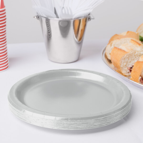 A stack of Creative Converting Shimmering Silver paper plates on a table next to a bucket of food.
