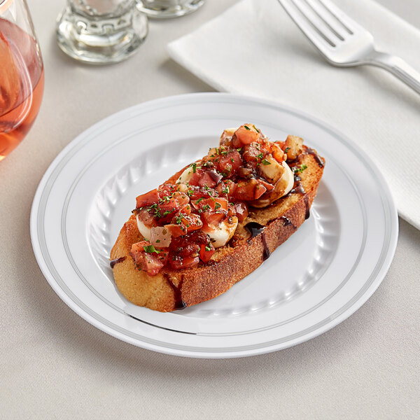 A Fineline white plastic plate with silver bands holding a piece of bread and a glass of wine on a table.