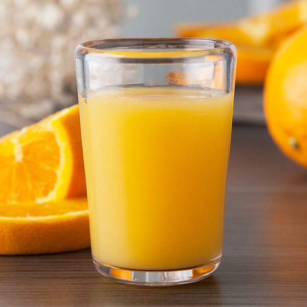 A close up of a Carlisle clear Tritan plastic juice glass filled with orange juice and a slice of orange.