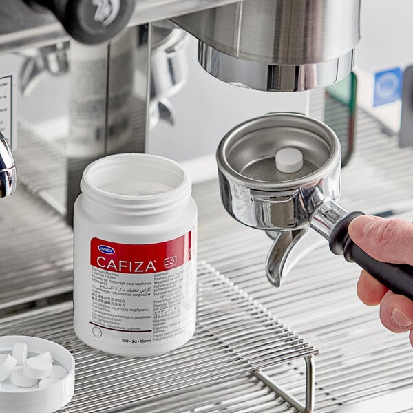 A container of Urnex Cafiza espresso machine cleaning tablets next to an espresso machine portafilter with a tablet inside.