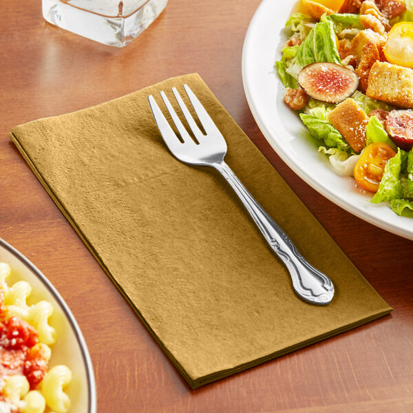 A Hoffmaster Glittering Gold paper dinner napkin with a fork on a table next to a salad plate.