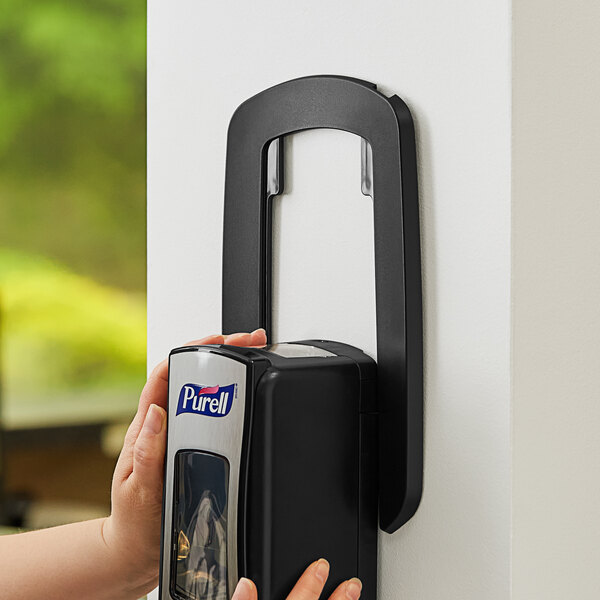 A hand using a black GOJO True Fit wall plate to hold a soap dispenser.