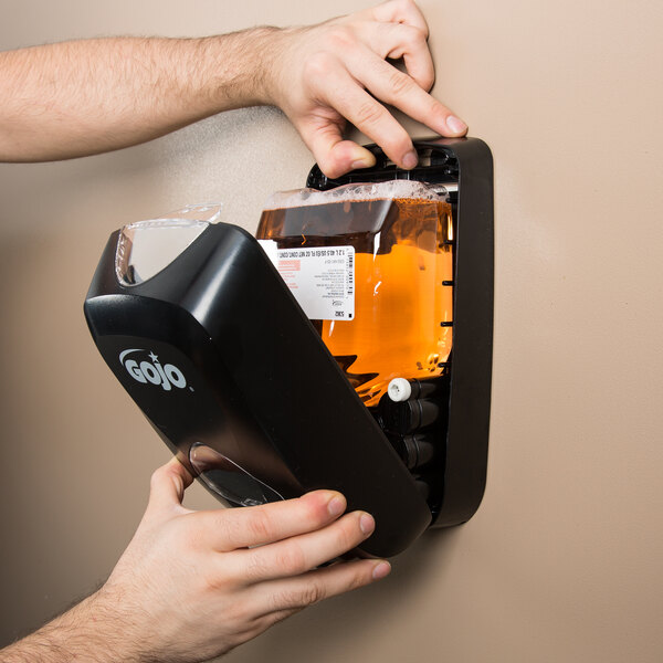 A hand holding a black GOJO TFX soap dispenser with a bottle of GOJO Fresh Fruit Foaming Antibacterial Hand Soap
