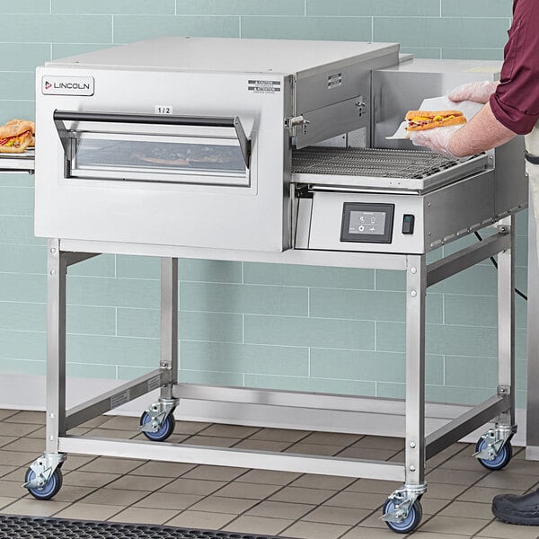 A man using a Lincoln stainless steel equipment stand to hold a pizza in a conveyor oven.