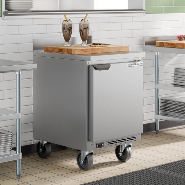 A stainless steel single door worktop freezer on wheels with a wooden cutting board on top.
