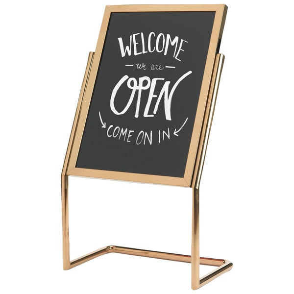 A black and gold Aarco double pedestal sign with white writing that says "Welcome to the Open" on it.