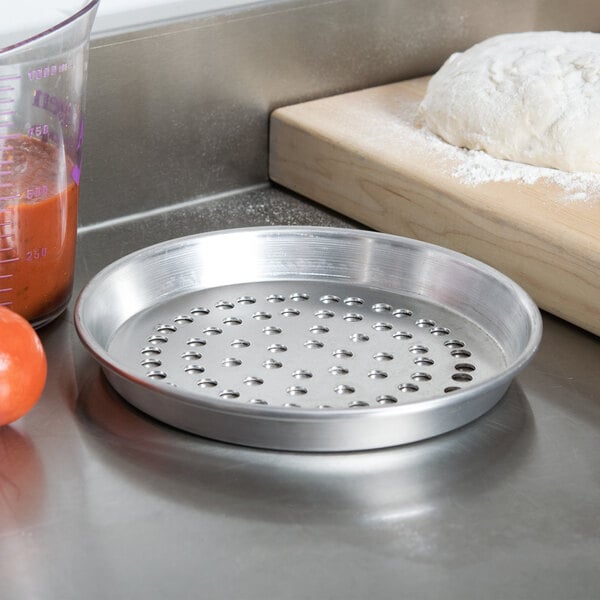 An American Metalcraft Super Perforated Deep Dish Pizza Pan with tomatoes, dough, and a measuring cup of liquid.