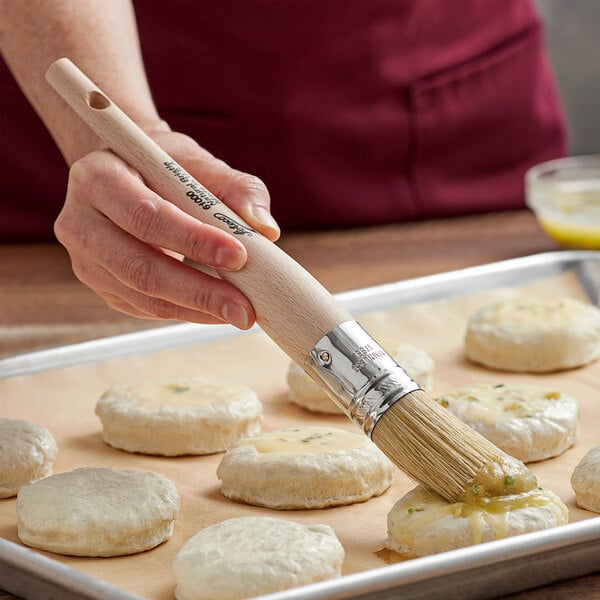 A person using an Ateco round boar bristle brush to baste a round white dough