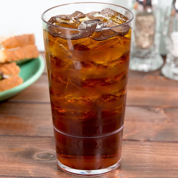 A close-up of a GET Clear SAN Plastic Tumbler filled with brown liquid and ice on a table.