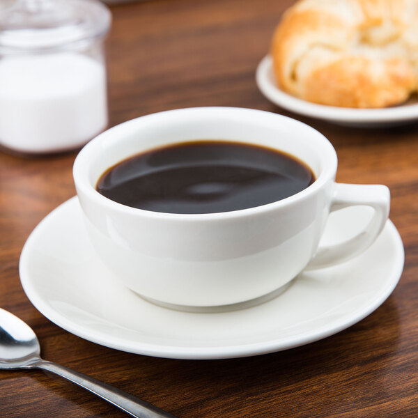 A CAC ivory china saucer with a cup of coffee on a table next to a croissant and a spoon.