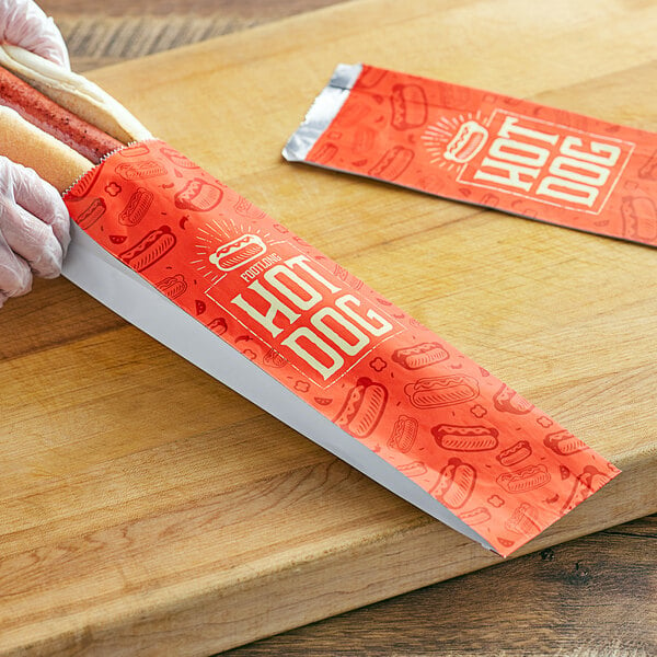 A person holding a footlong hot dog in a red and white printed paper wrapper.