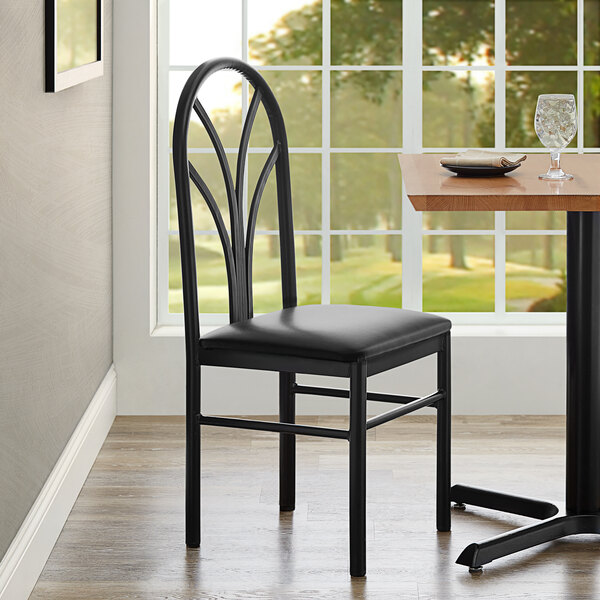 A black Lancaster Table & Seating chair with a black vinyl seat next to a dining table with a place setting.
