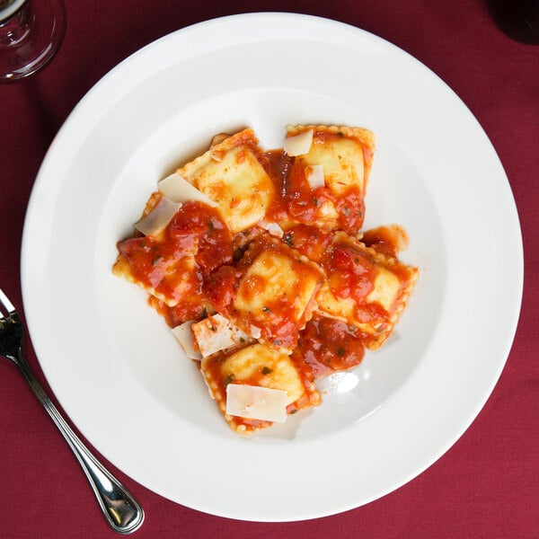 A Tuxton eggshell china pasta bowl filled with ravioli in sauce on a table with a glass of wine and a fork.
