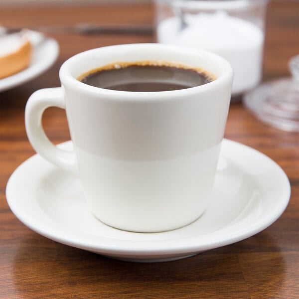 A Tuxton eggshell tall china cup filled with brown liquid on a saucer.