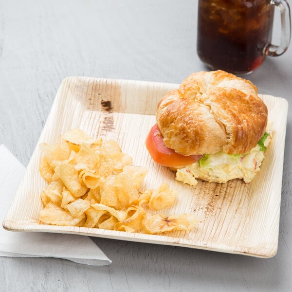 A croissant sandwich and chips on an EcoChoice palm leaf plate with a drink.