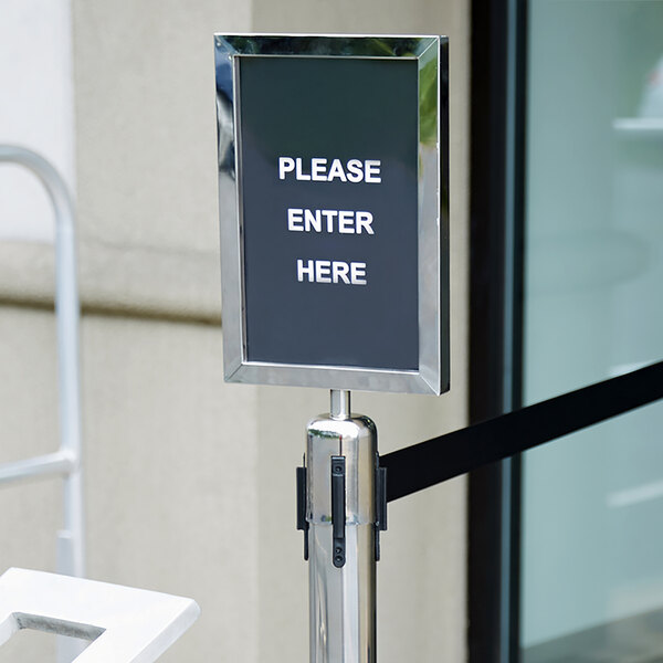 A Lancaster Table & Seating crowd control stanchion with a clear sign that says "Please Enter Here" on a pole.
