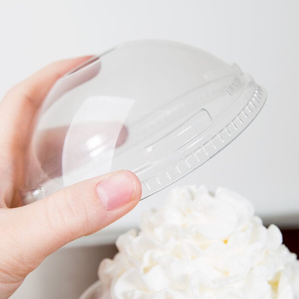 A hand holding a Solo clear plastic dome lid over whipped cream on a clear plastic container.