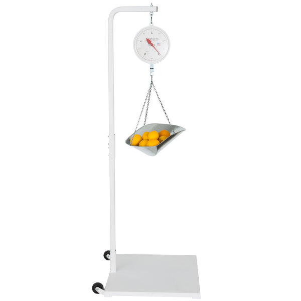 A Cardinal Detecto hanging scale with a metal scoop full of oranges and a basket of oranges.