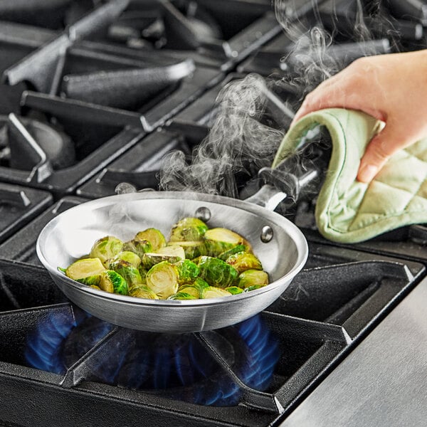 A hand using a potholder to pour brussels sprouts from a pot into a Vollrath aluminum fry pan with a black silicone handle.