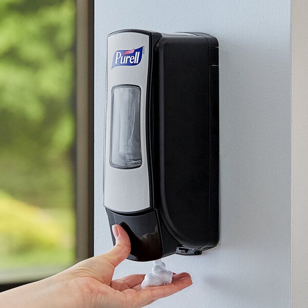 A person using a Purell brushed chrome and black hand sanitizer dispenser on a wall.