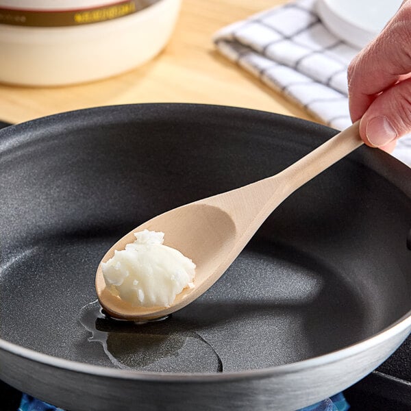 A person using a wooden spoon to stir white coconut oil in a pan.
