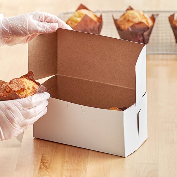 A person in gloves holding a white bakery box of muffins.