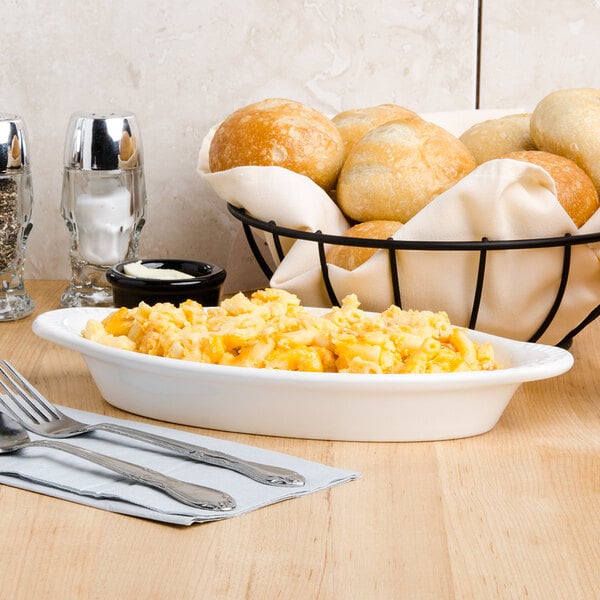 A white Tuxton oval bowl filled with macaroni and cheese on a table with a napkin, fork, and spoon.