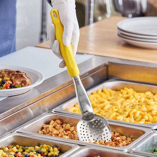 A person using a Vollrath Jacob's Pride heavy-duty perforated basting spoon with a yellow handle to serve food.
