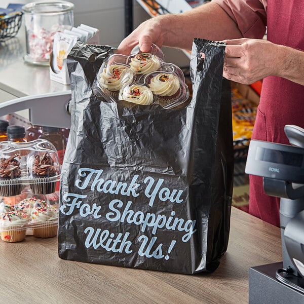 A person holding a black plastic Choice T-shirt bag with cupcakes inside.