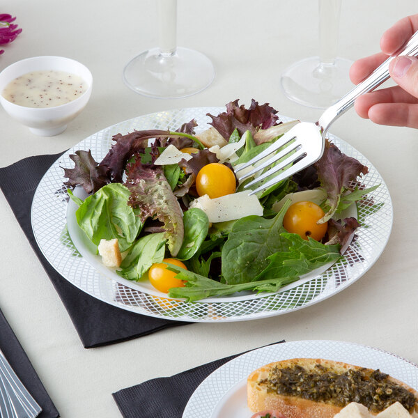 A hand holding a fork with a salad on a white and silver Visions plastic plate.