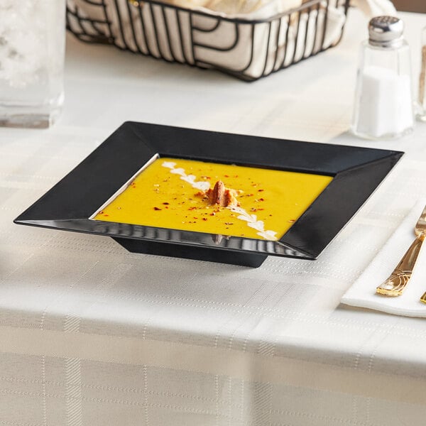 A black square plastic bowl with yellow soup in it on a black square plate.