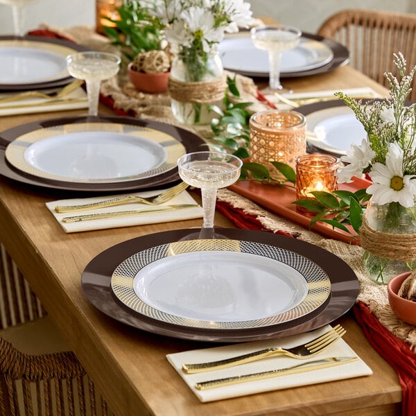 A wooden dining table is elegantly set with patterned plates, clear plastic champagne glasses, gold-colored cutlery, floral centerpieces, and decorative candles.