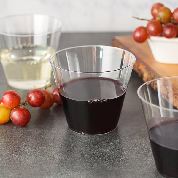 Two glasses of red wine and a glass of white wine in hard plastic cups with grapes in background
